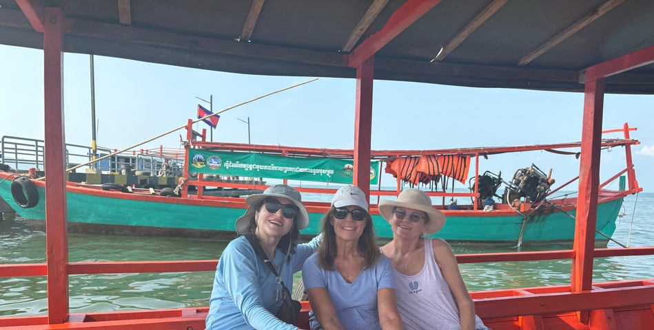       Three women sitting on a boat with a colorful fishing boat in the background.
  
