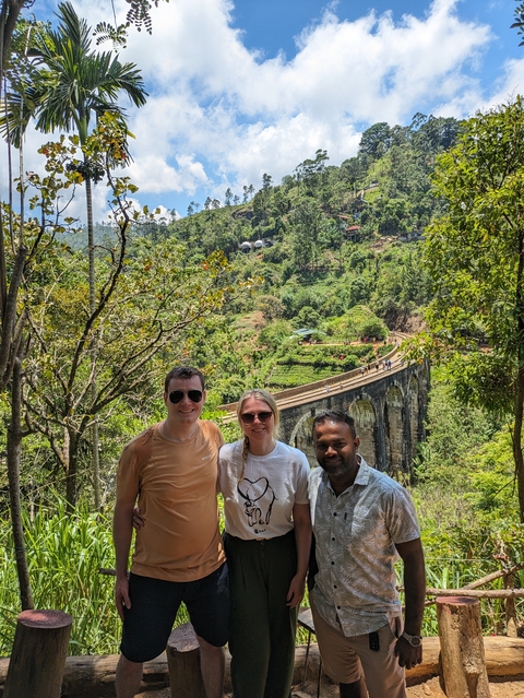 Three people posing in front of a railway bridge and lush landscape.