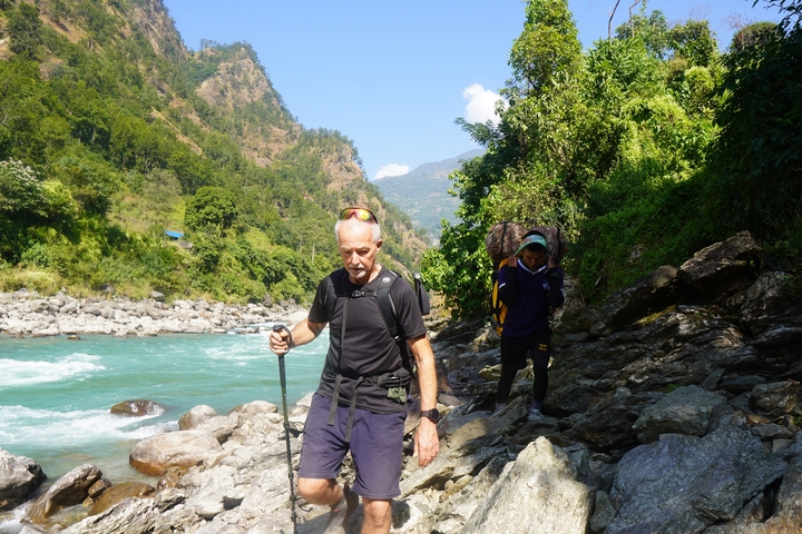      Two people hiking on a rocky path near a river with mountains in the background.
  