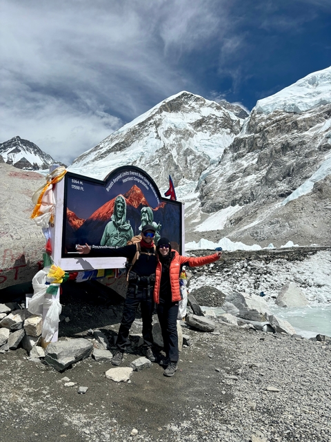 Two people at a mountain base camp with a sign and flags.