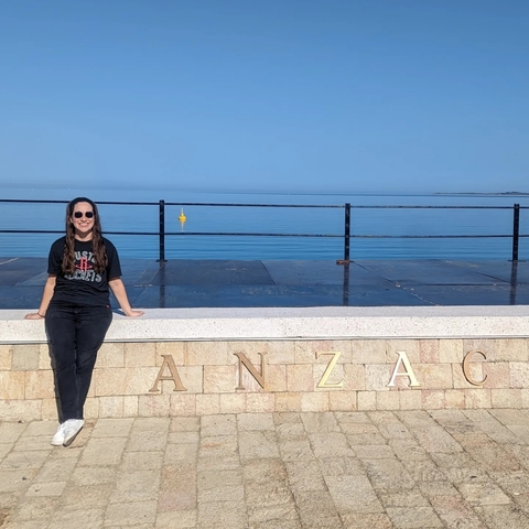 A person sitting by a stone railing with 'ANZAC' spelled out, overlooking a calm sea with a clear blue sky.