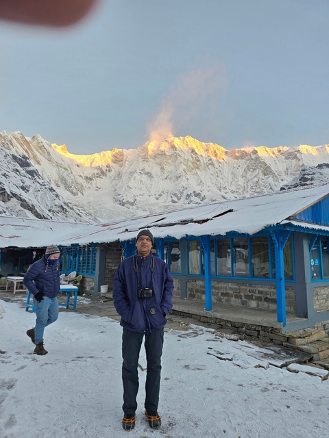 Man posing in front of a mountain lodge with sunlit peaks.
