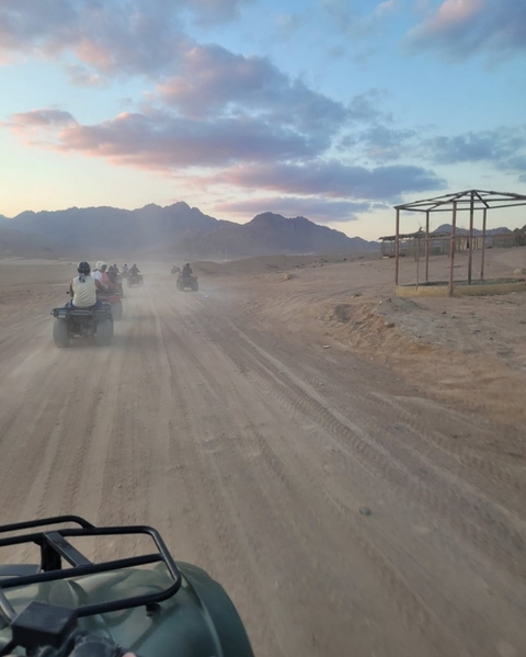 People riding ATVs on a dusty desert road with mountains in the background.