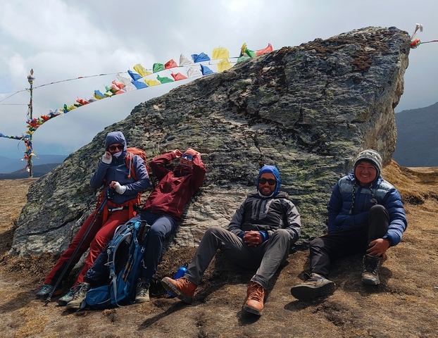       Hikers resting against a large rock with prayer flags above.
  