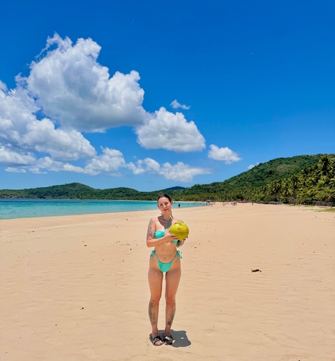       Woman holding a coconut on a sandy beach.
  