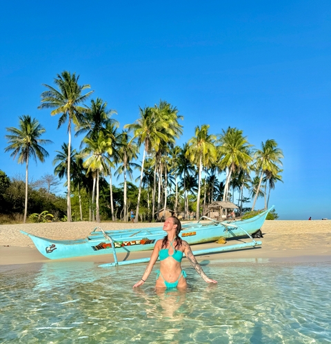 Woman sitting on a beach near a boat with palm trees in the background.