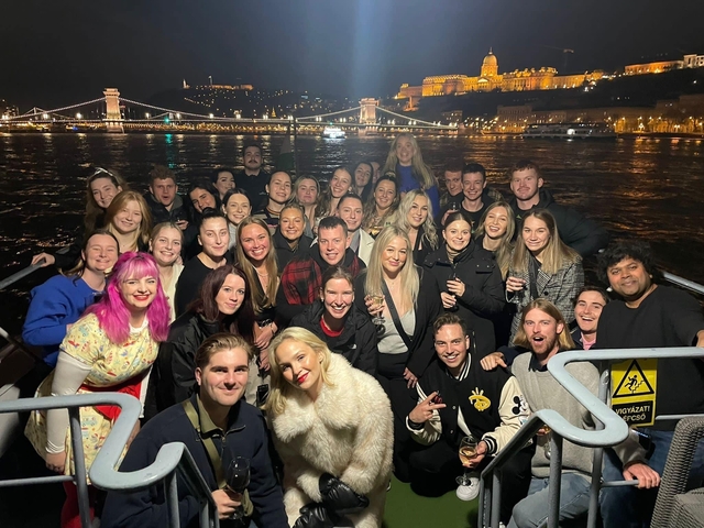 Large group on a boat at night with the Budapest skyline in the background.