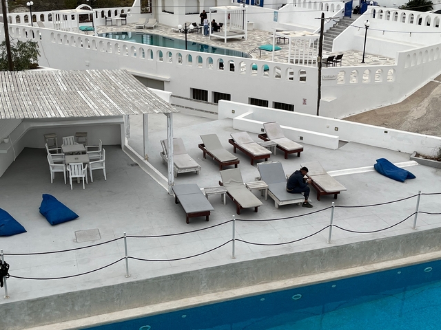 Person sitting alone on a terrace by a pool in a white-washed building setting.