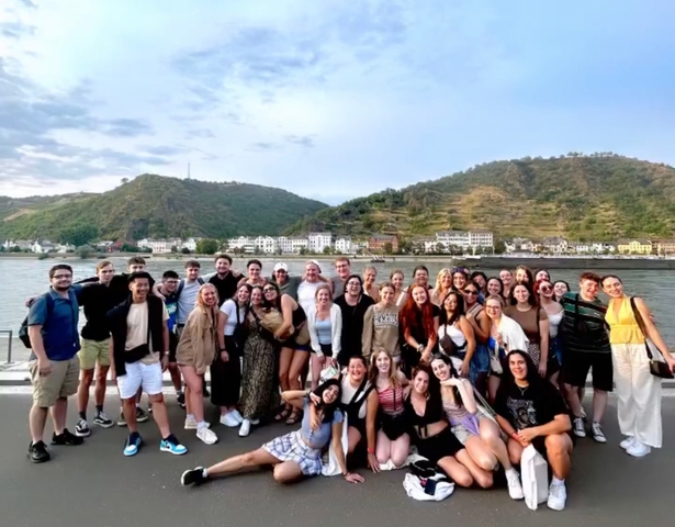 A large group of people posing on a boat with the river and hills in the background.