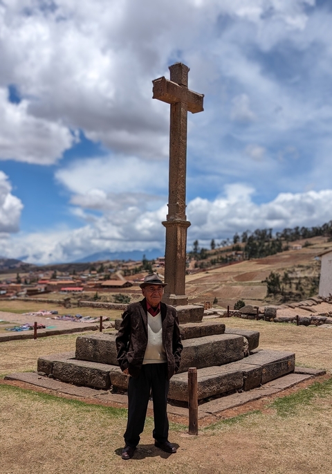 An elderly man standing in front of a historical pillar in a rural setting.