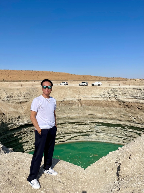 Person standing near a desert crater with vehicles in the background.