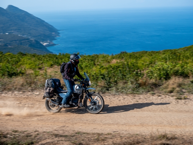       Person riding a motorcycle on a dirt road with sea in the background.
  