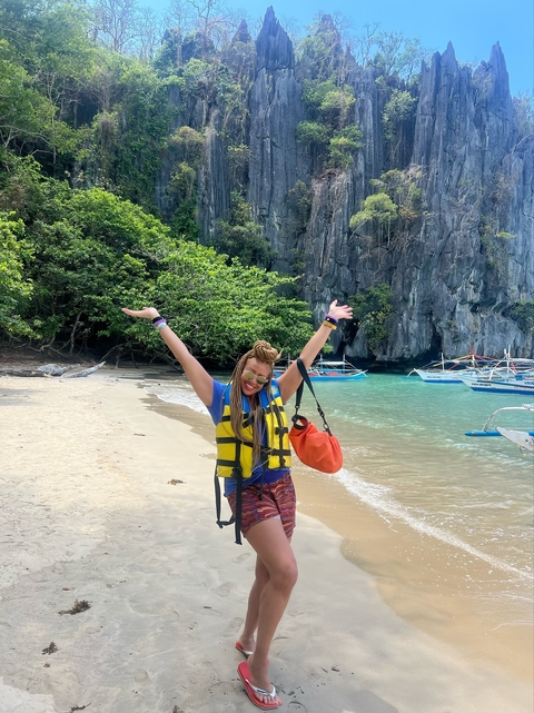       Person in a life jacket smiling on a beach with boats and cliffs.
  