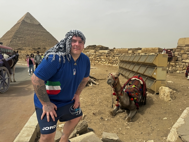 Man posing near a camel and pyramid.