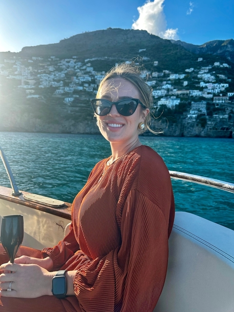 Woman in sunglasses sitting on a boat with scenic water and mountain backdrop.