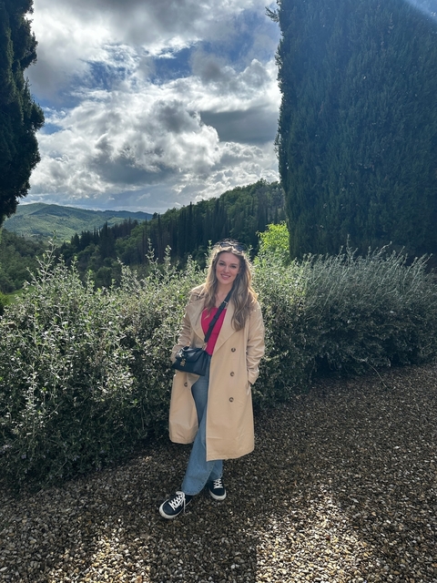 Woman posing in a trench coat amidst lush greenery and hills.