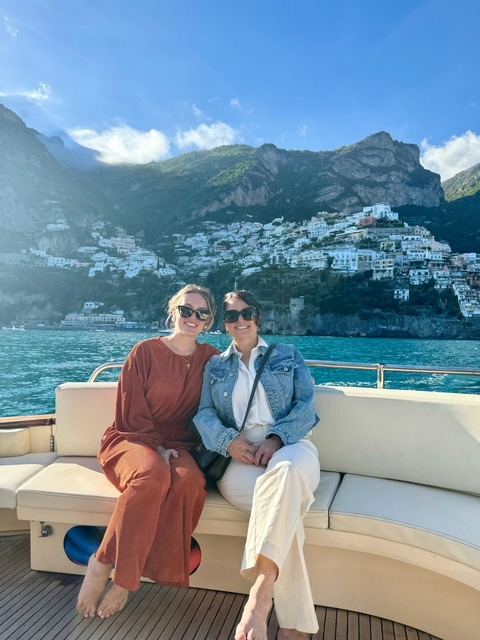 Two women on a boat in front of a coastal town with buildings built on the cliff.