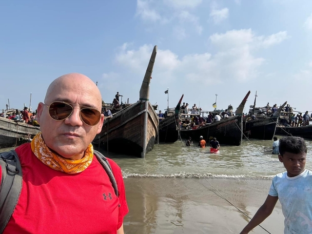 Man with sunglasses standing on a beach with boats and people in the background.