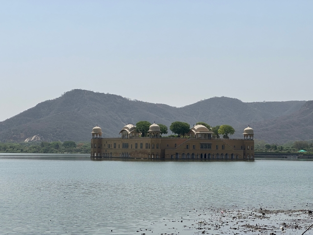       Jal Mahal in the middle of a lake with mountains in the background.
  