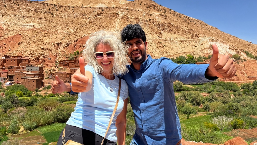       Two people giving thumbs up with a village and hills backdrop.
  