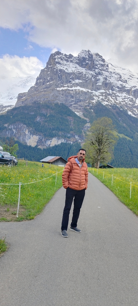       A person in a puffer jacket walking on a path in a mountainous landscape with trees and small buildings.
  