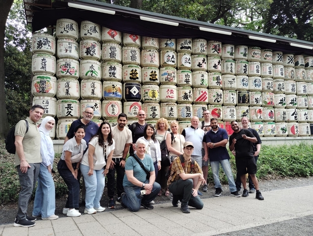       A group of people posing in front of stacked barrels with Japanese labels.
  
