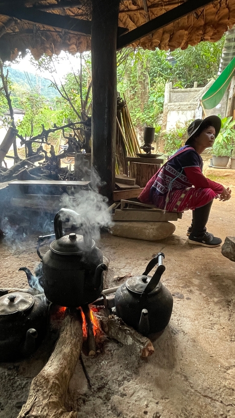       Person sitting inside a traditional wooden cabin with steaming kettles.
  
