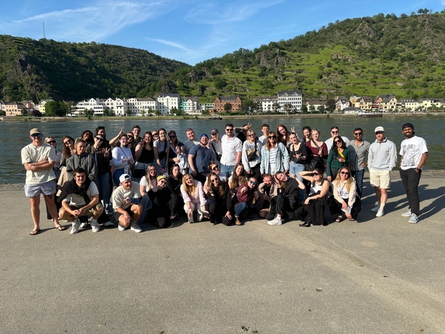Group of people by a river with hills and colorful buildings in the background.