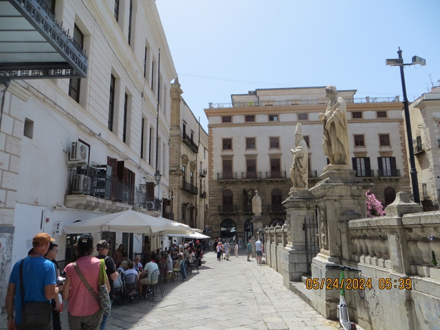       Street view with dining areas and statues in an Italian city.
  