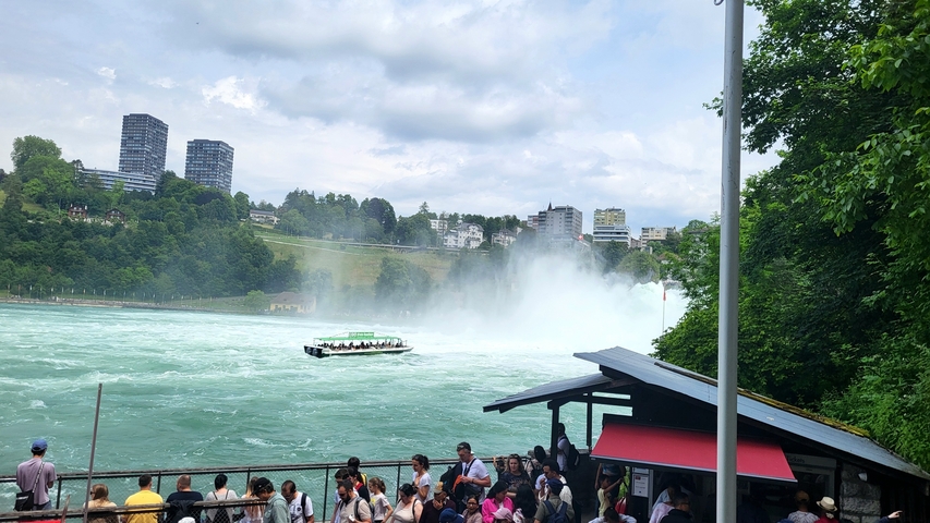 Waterfalls with tourists by a riverbank and urban backdrop.