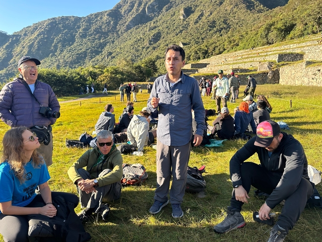 A man standing and addressing a group of people sitting on grass near ancient ruins.