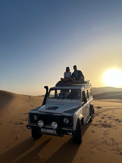 Two people sitting on top of a vehicle in the desert during sunset.