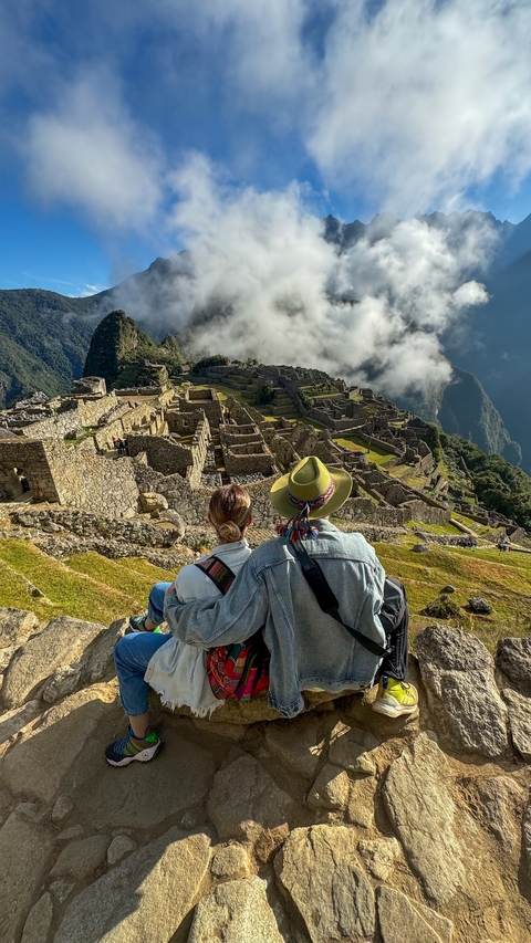       Couple sitting and admiring Machu Picchu ruins with clouds around.
  