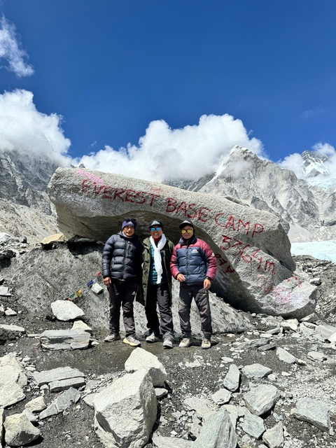 Three people standing in front of a large rock with Everest Base Camp inscription, snowy mountains in the background.