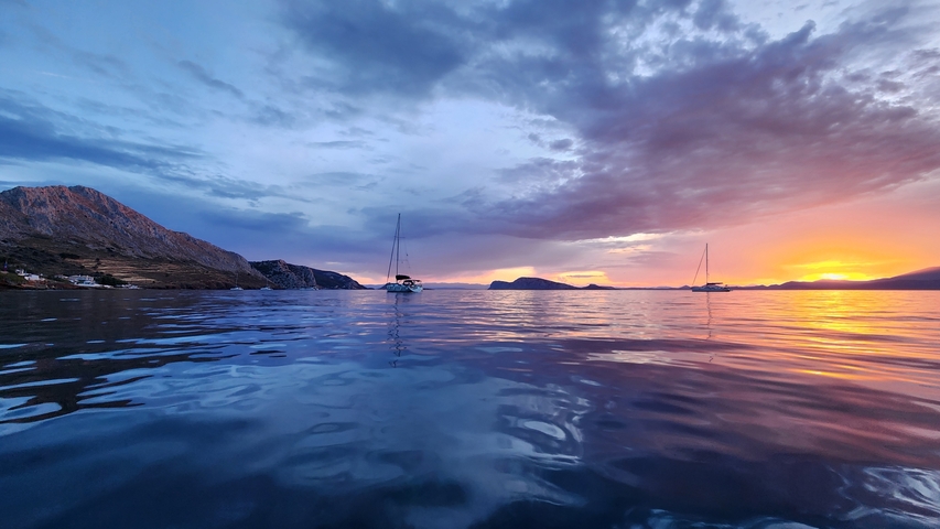       Sunset over the sea with sailboats and distant mountains.
  