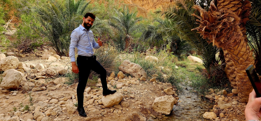 Person smiling in front of palm trees and rocky landscape.