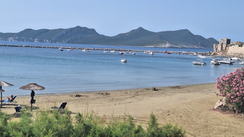       Beach scene with sun umbrellas, chairs, a rocky hill, and a historic fortress.
  