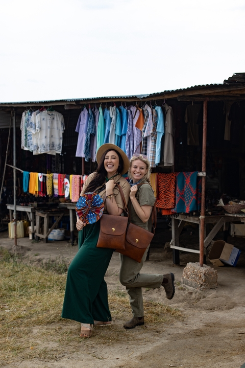Two women posing in front of a market with colorful cloths and items.