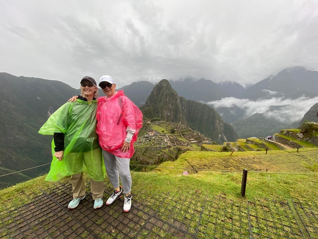       Two people in bright ponchos overlooking Machu Picchu on a cloudy day.
  
