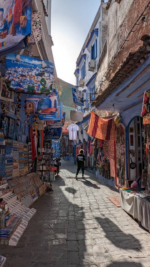       Narrow street in a market with hanging textiles and signs.
  