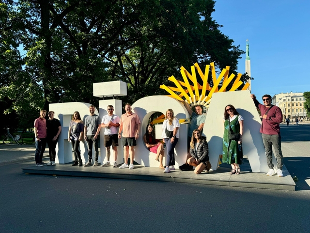 Group posing with 'Riga' lettering and cityscape background.