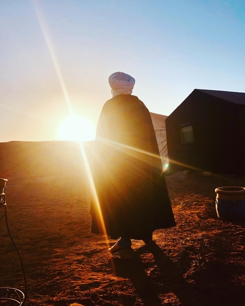 A silhouette of a person standing by tents with the sun setting behind them.