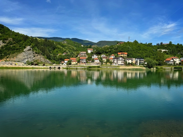 Serene lake with reflection of houses and forested hills.