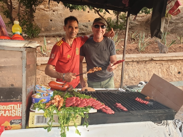       Two men grilling meat at an outdoor food stall.
  