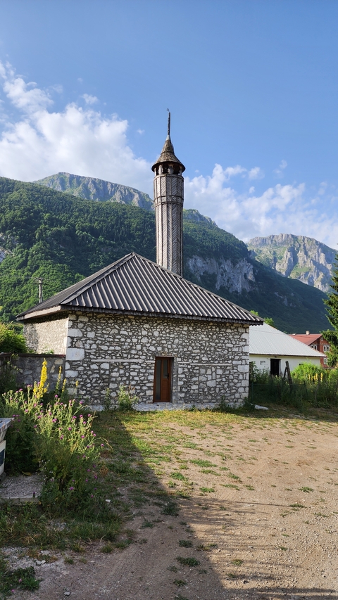 Small stone building against a backdrop of forested mountains.