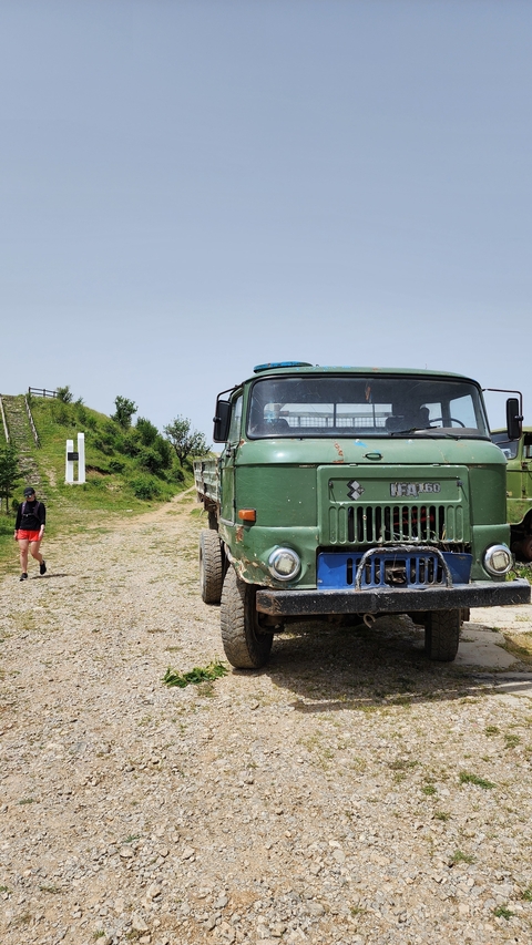 Rustic green truck parked on a dirt path with a person walking nearby.