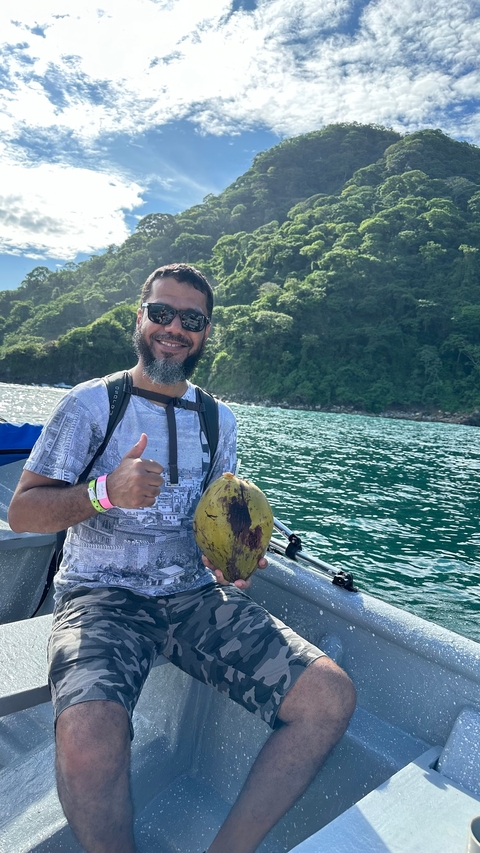 Smiling man holding a coconut by water with greenery in the background.
