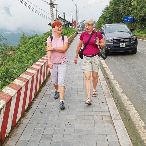       Two people walking along a sidewalk with a vehicle in the background.
  
