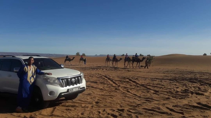 A man stands next to an SUV with camels in the desert.