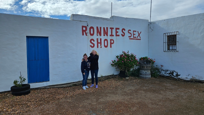       Two people standing in front of a building with 'Ronnies Sex Shop' painted on it.
  